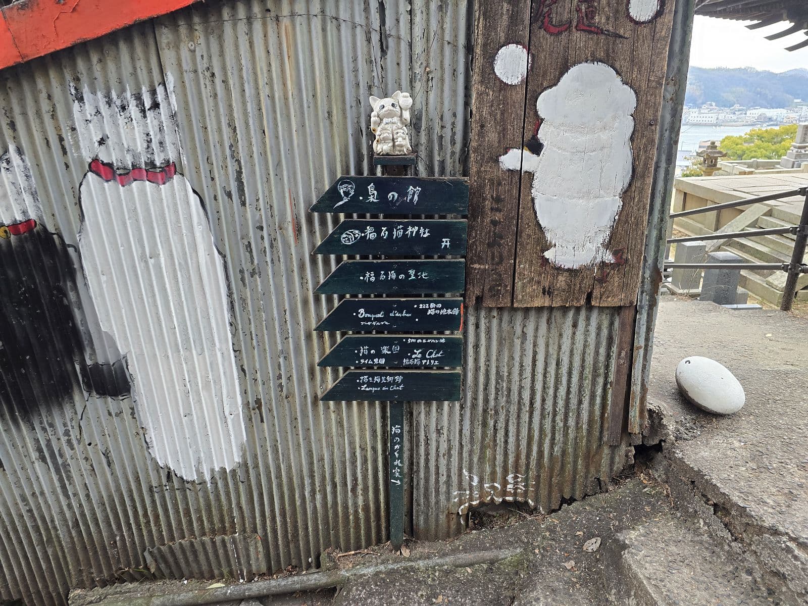 猫の細道・福石猫神社の道しるべ——トタン板の壁に黒板看板と白い猫のシルエットアート